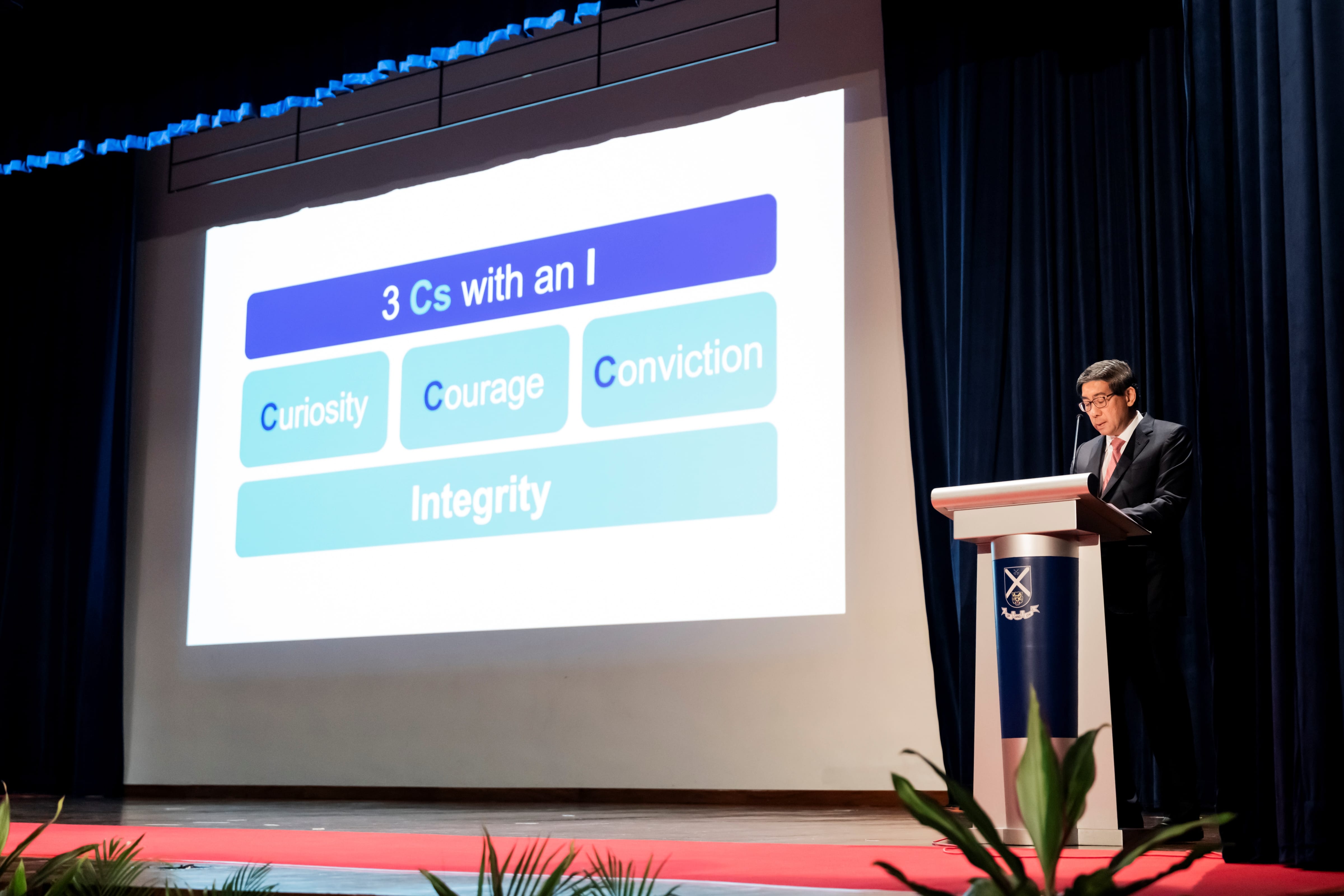 Guest of Honour, Dr Yeo, stands at a podium next to a presentation slide displaying a diagram titled "3 Cs with an I: Curiosity, Courage, Conviction, and Integrity."